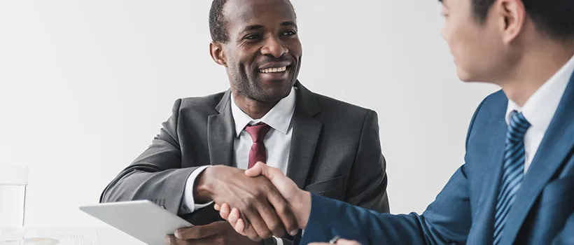 A photo of 2 men shaking hands over a tablet being held by one of the individuals.