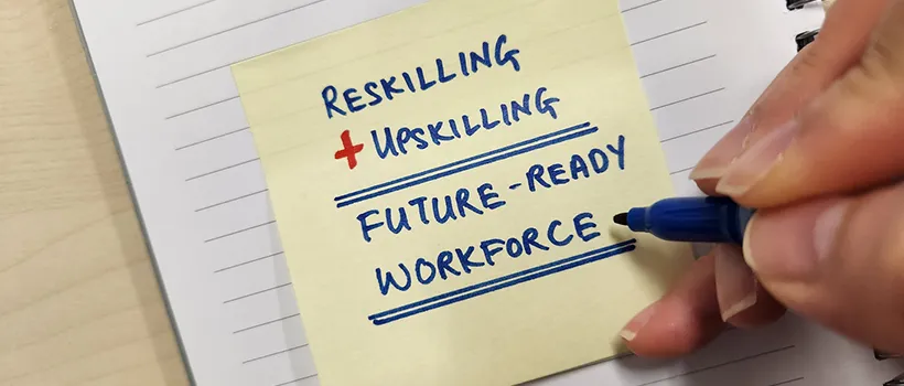 A person writing a note on a post-it note that reads "Reskilling + Upskilling. Future-ready workforce"