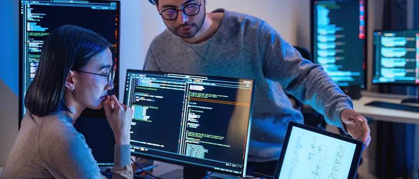 A photo of a woman looking at code on a monitor with a male co-worker looking over her monitor.